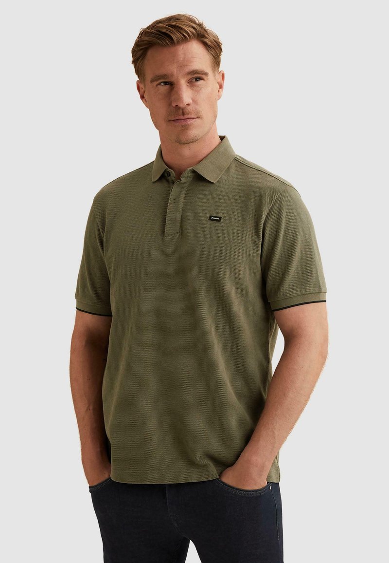 Homme aux cheveux châtain clair portant un polo vert olive à manches courtes et un pantalon foncé, mains dans les poches, regardant légèrement vers la gauche.