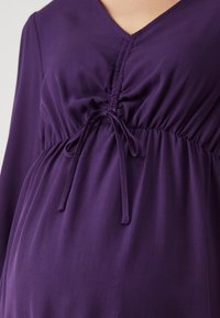 Närbild av en lila långärmad klänning med V-ringning och veckad front som är knuten med en rosett vid midjan.