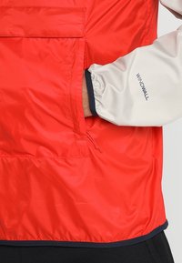 Veste coupe-vent rouge avec une texture lisse et légère. Elle présente une manche grise avec un poignet froncé et une poche visible à l'avant.