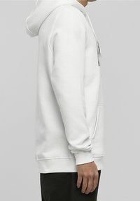 Sudadera con capucha de algodón blanco con bolsillo frontal, capucha con cordón y puños acanalados, usada por una persona vista de perfil sobre un fondo gris.