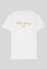 Camiseta de algodón blanca con texto dorado que dice "Palm Spring" con "California Love Eighty Five" debajo, de corte relajado y diseño de cuello redondo.