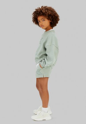 Barn med krøllet hår iført lysegrøn oversized sweatshirt og matchende shorts, stående med hænderne i lommerne mod en ensfarvet baggrund.