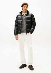 Άνδρας φορώντας μαύρο φουσκωτό μπουφάν, γκρι πουλόβερ με καλωδίωση, άσπρα παντελόνια και μαύρα loafers, στέκεται μπροστά σε άσπρο φόντο.