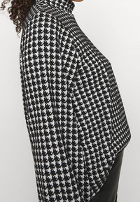 Haut à motif pied-de-coq noir et blanc avec un col montant, présentant des manches longues et une coupe ample, fabriqué en tissu tricoté texturé.