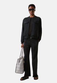 Schwarze Bomberjacke mit Reißverschluss und Fronttaschen, kombiniert mit passenden Hosen. Graue, wattierte Tote-Bag und schwarze Sandalen vervollständigen den Look.