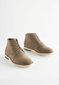 Stivali alla caviglia in suede marrone con chiusura a lacci, tomaia texture, suola in gomma bianca e accenti neri a contrasto sul tacco.