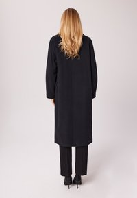 Długi czarny płaszcz z wełny syntetycznej, o prostym kroju, z długimi rękawami i bocznymi kieszeniami, w zestawieniu z czarnymi zwężającymi się spodniami i szpilkami.