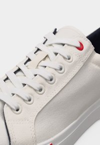 Λευκό καμβαδένιο αθλητικό παπούτσι με λευκά κορδόνια, λευκές τρύπες για τα κορδόνια, μια κόκκινη τρύπα στην κορυφή και λευκή σόλα σε μονόχρωμο φόντο.