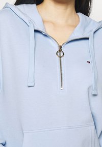 Sudadera con capucha azul claro, con media cremallera, que presenta un tirador de cremallera dorado, puños acanalados y un bolsillo en la parte frontal. Logo exhibido en el pecho.