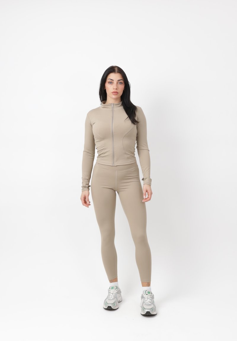 Femme debout devant un mur blanc portant une veste beige ajustée avec fermeture éclair, des leggings assortis et des baskets blanches de sport.
