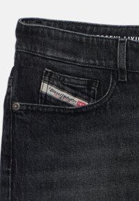 Les jeans en denim noir présentent un design classique à cinq poches, des accessoires en métal argenté et une étiquette de marque visible dans la poche avant.