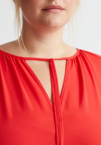 Rote Bluse mit einem V-Ausschnitt und einem Schlüsselloch-Ausschnitt, weichem Stoff und gerafftem Detail an den Schultern. Glatte Textur, figurbetontes Design.