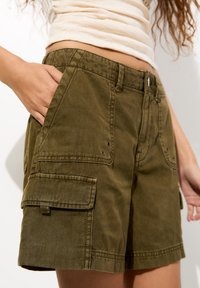 Pantaloni cargo verde oliva realizzati in cotone, con tasche anteriori, alette laterali e chiusura con bottone. Presentano dettagli di cucitura e una vestibilità comoda.