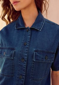 Camisa de denim en azul oscuro, con cuello, mangas cortas, dos bolsillos delanteros y seis botones negros en la parte frontal.