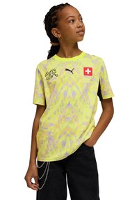 T-shirt pour jeunes jaune à motif, col rond, arborant un logo du drapeau suisse et une marque Puma. Tissu texturé avec manches courtes.