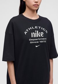 Νεαρός/Νεαρή φοράει μαύρο φαρδύ μπλουζάκι Nike με λευκό κείμενο που λέει "ATHLETICS nike Department Since 1972" και μικρό λογότυπο Nike.