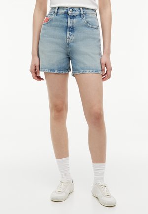 Persona que lleva shorts vaqueros de tiro alto de color azul claro, calcetines blancos, zapatillas blancas y una camiseta blanca, de pie frente a un fondo blanco.