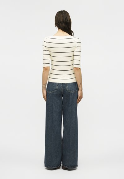 Γυναίκα που στέκεται γυρισμένη από την πλάτη, φοράει λευκό εφαρμοστό τοπ με μαύρες οριζόντιες ρίγες και σκουρόχρωμο μπλε τζιν με φαρδιά μπατζάκια.