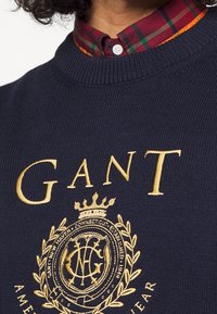 Granatowy sweter z wyhaftowanym złotym napisem "GANT" oraz logo z koroną i wieńcem laurowym.