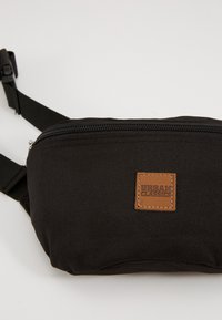 Marsupio nero realizzato in tessuto resistente, dotato di una zip prominente, di una tracolla regolabile e di un'etichetta marrone cucita con la scritta "Urban Classics".