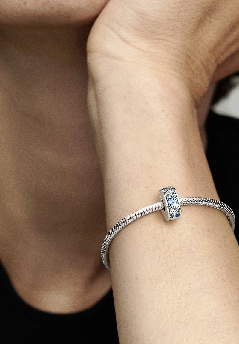 Srebrna zapestnica z teksturiranim, tkano obliko. Ima dekorativen obesek z modrimi in zelenimi poudarki. Gladka površina, eleganten oblik.