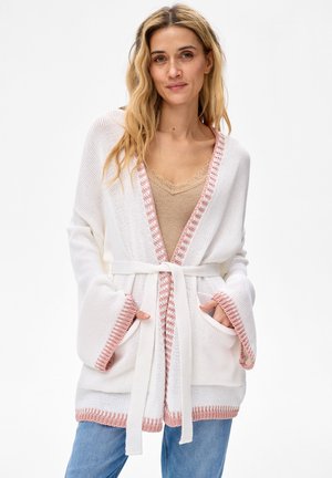 Vrouw draagt een witte gebreide cardigan met roze afwerking en riem over een beige top met kanten rand en een blauwe spijkerbroek, handen in de zakken, neutrale achtergrond.