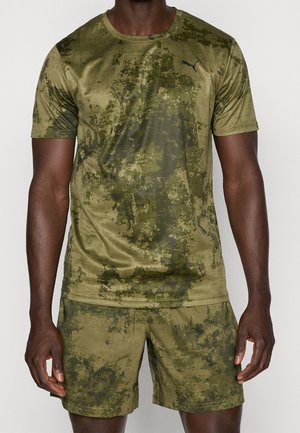 Mann trägt olivgrünes Puma-T-Shirt mit Camouflage-Muster und passende Shorts, steht vor schlichtem hellem Hintergrund.