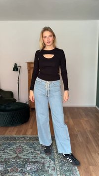 Νέα γυναίκα που στέκεται μέσα, φοράει μαύρο μακρυμάνικο τοπ με άνοιγμα, ανοιχτό μπλε τζιν με κορδόνι και μαύρα αθλητικά παπούτσια Adidas.