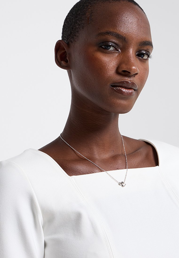 Collier en argent avec un petit pendentif sur un mannequin, associé à une robe blanche à encolure carrée et à la texture lisse.