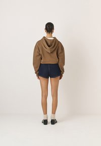 Barna, oversized kapucnis pulóver elülső zsebbel, sötét farmer sorttal párosítva. A modellen fekete cipő és krémszínű zoknik vannak. Egyszínű háttér.