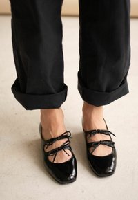 Ballerine in vernice nera con punta quadrata, caratterizzate da lati aperti e due fiocchi nella parte anteriore. Indossate con pantaloni neri a tinta unita e orlo arrotolato.