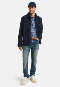 Marine denimjack over een blauw bloemenoverhemd, lichtblauwe jeans en witte sneakers. Heeft knopen, een riem en een donkere pet.