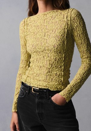 Femme portant un haut moulant jaune à manches longues en dentelle florale, rentré dans un jean noir avec une ceinture noire, main dans la poche.