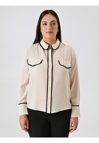 Frau mit langen dunklen Haaren, die eine cremefarbene Bluse mit schwarzer Borte an Kragen, Taschen, Manschetten und Knopfleiste trägt, kombiniert mit schwarzen Hosen.