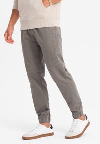 Graue, schmal zulaufende Hose mit glatter Textur, ausgestattet mit einem elastischen Bund und Bündchen, kombiniert mit weißen Sneakers und einem beigen Oberteil.