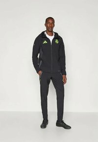 Zwarte sportjasje en broekset met groene accenten en logo. Jas heeft een ritssluiting en capuchon. Soepele textuur, atletische pasvorm.