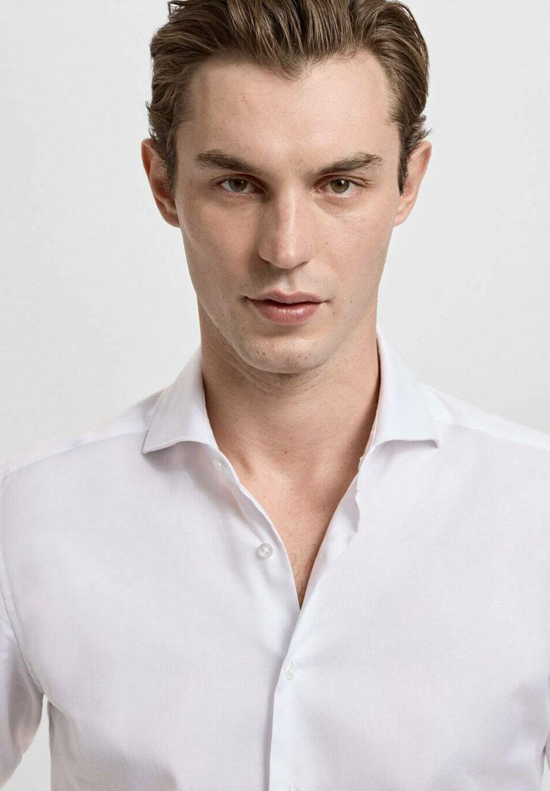 Chemise blanche à boutons avec col classique, patte de boutonnage apparente et texture lisse, portée par un mannequin sur un fond neutre.