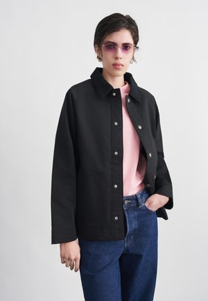 Person wearing purple-tinted glasses, black buttoned jacket with large collar, pink shirt, and blue jeans, standing against a plain background.