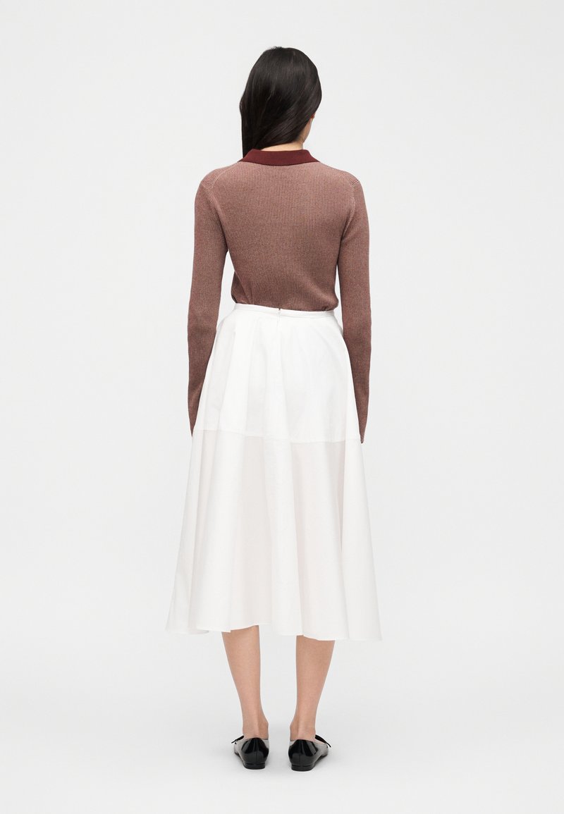 Donna con capelli lunghi e scuri che indossa una maglia marrone a maniche lunghe e una gonna midi bianca a vita alta, vista di spalle, su uno sfondo semplice.