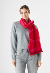Roter Schal mit pinkem Muster, aus weichem Stoff, um den Hals gewickelt. Grauer Pullover mit geripptem Saum und hellgraue Jeans ergänzen das Outfit.