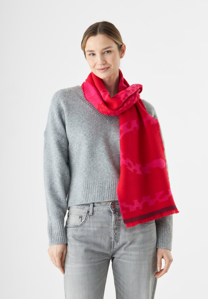 Roter Schal mit pinkem Muster, aus weichem Stoff, um den Hals gewickelt. Grauer Pullover mit geripptem Saum und hellgraue Jeans ergänzen das Outfit.