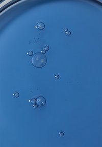 Niebieska powierzchnia cieczy z różnymi przezroczystymi bąbelkami o różnych rozmiarach, odbijającymi światło i tworzącymi teksturowany wygląd.