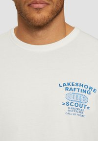 Biała bawełniana koszulka z niebieskim nadrukiem tekstu "LAKESHORE RAFTING" oraz "SCOUT" z ilustracją łodzi i detalami poniżej.