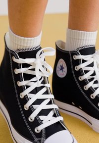 Svarta canvas sneakers med hög sko har vita snören, en gummikapsel vid tån, kontrasterande sömmar och en cirkulär logotyp på den yttre sidan.