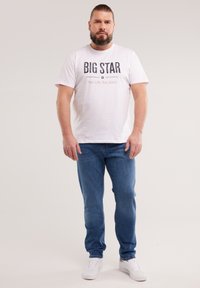 Uomo con barba e capelli legati indossa una maglietta bianca con la scritta "BIG STAR", jeans blu e sneaker bianche, in piedi davanti a uno sfondo uniforme.