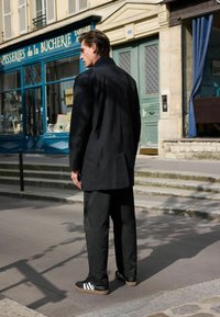 Geacă lungă neagră dintr-un material texturat, asortată cu pantaloni negri largi și adidași negri cu accente albe. Cadru urban.