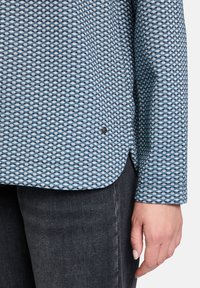 Langarmhemd in Blau mit geometrischem Muster, aus strukturiertem Stoff gefertigt, mit abgerundetem Saum und einem kleinen dekorativen Knopfdetail.