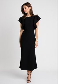 Czarna, dopasowana sukienka z falbaniastymi rękawami i rozkloszowanym dołem. Gładka tekstura, do kolan, zestawiona z czarnymi sandałami na obcasie i zegarkiem na nadgarstku.