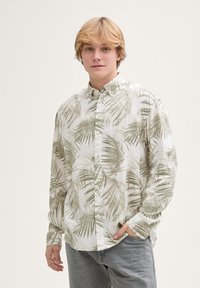 Camicia bianca a maniche lunghe con stampa di foglie di palma verdi, colletto con bottoni e vestibilità rilassata. Abbinata a jeans azzurri chiari.