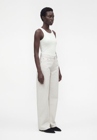 Femme debout en haut blanc sans manches, pantalon large taille haute couleur écru et chaussures pointues noires sur fond blanc uni.